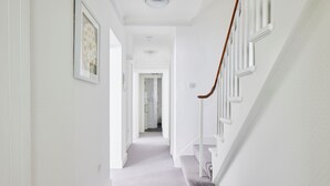 Un couloir étroit et blanc, doté d’un sol recouvert de moquette, d’un escalier avec une rampe en bois et d’un tableau encadré accroché au mur.