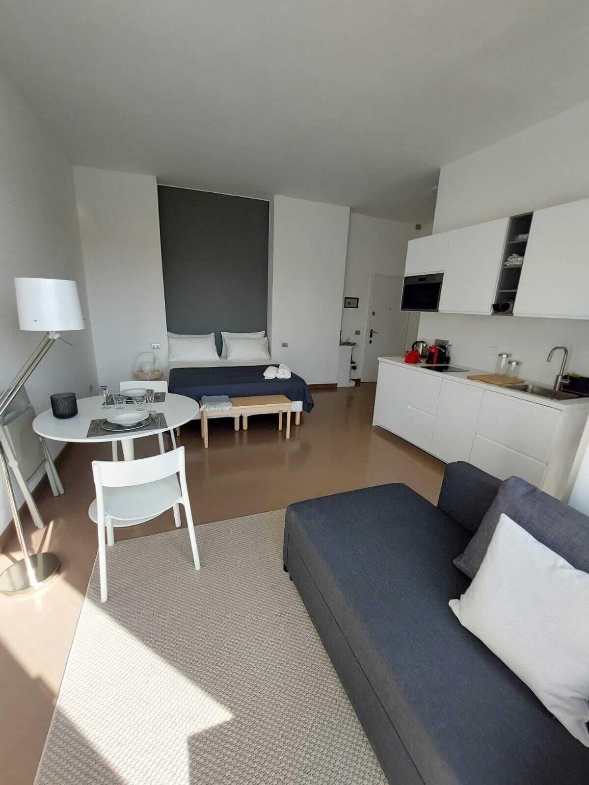 Ein modernes, helles Zimmer mit Bett, einer Couch, einem Esstisch mit Stühlen, einer Küchenzeile und einem großen Fenster.