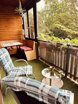 Un espace extérieur aménagé, abrité, avec un banc en bois, deux fauteuils, une petite table et une plante suspendue.