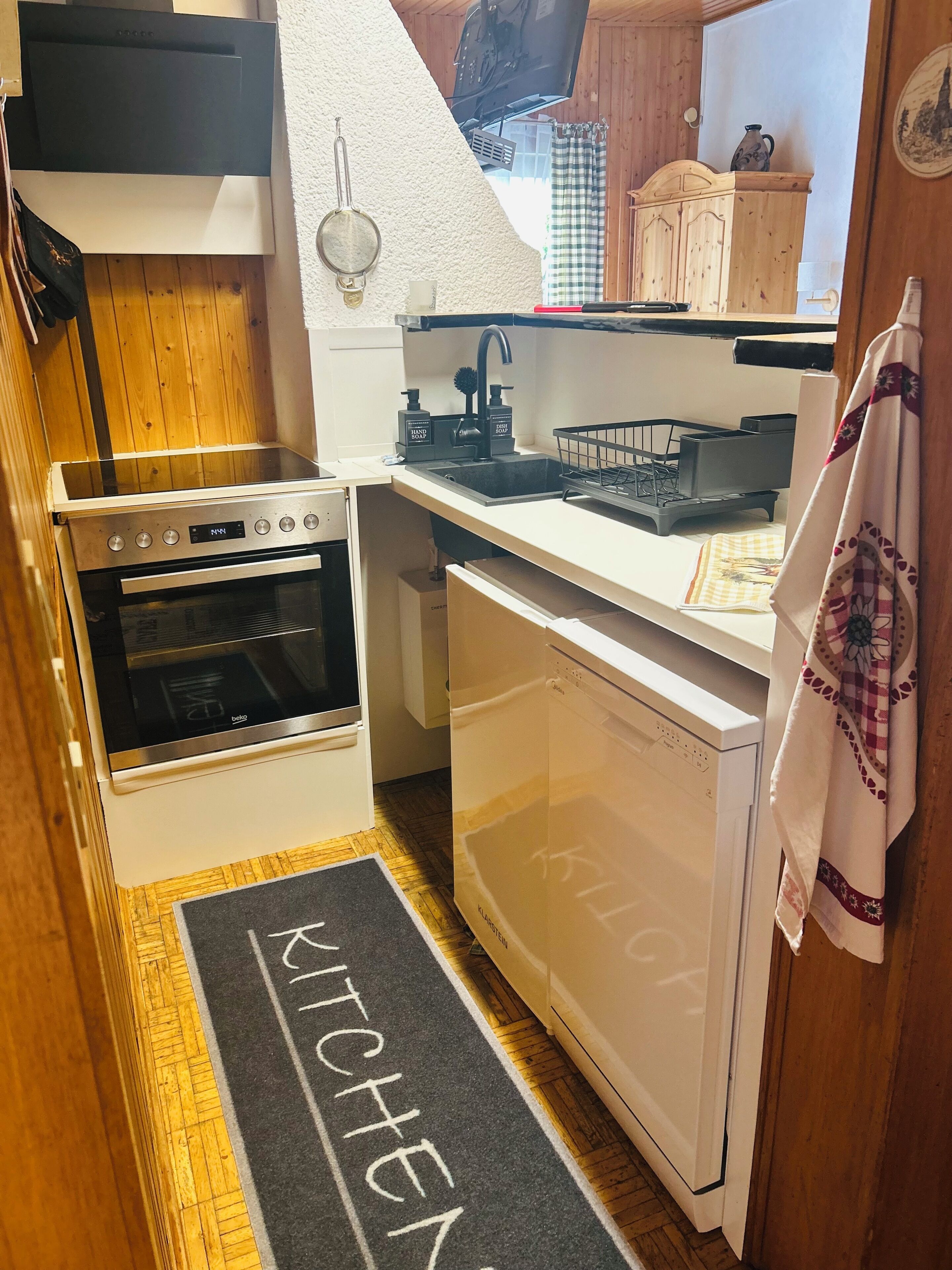 Cocina compacta con horno integrado, lavavajillas y un lavamanos con grifería moderna.