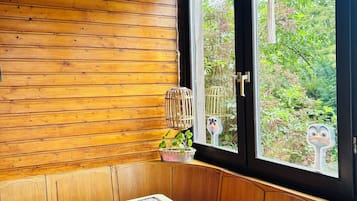 Une pièce en bois avec un banc, une table recouverte d’une nappe, une fenêtre donnant sur un paysage verdoyant et une plante suspendue.