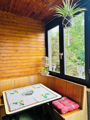 Une pièce en bois avec un banc, une table recouverte d’une nappe, une fenêtre donnant sur un paysage verdoyant et une plante suspendue.