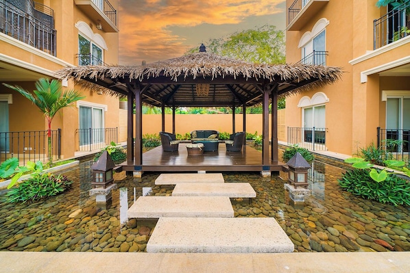 A covered outdoor seating area with a thatched roof, surrounded by a water feature and steps leading to a pool.