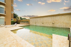 A swimming pool with a tiled deck, lounge chairs, and a building with balconies.
