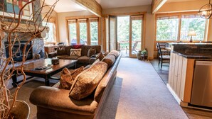 Ein geräumiges Wohnzimmer mit einer langen Couch, einem Couchtisch und einem Kamin. Es gibt eine Küchenzeile mit Holzausbau und einem Geschirrspüler. Große Fenster sorgen für natürliches Licht und Ausblicke ins Freie.