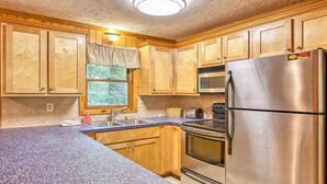 A kitchen with wooden cabinets, a stainless steel refrigerator, a microwave, a stove, and a window with a view of trees.