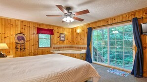 A wooden cabin bedroom with a bed, a ceiling fan, a window with a view of greenery, a bathtub, and a dreamcatcher on the wall.