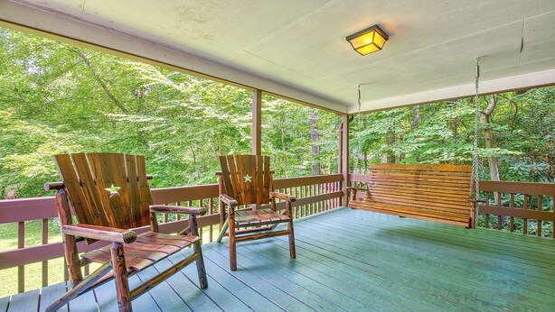 A covered porch with two Adirondack chairs and a bench, surrounded by lush greenery.
