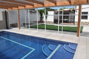 A swimming pool with a tiled deck, wooden beams, and a glass enclosure leading to a patio area with a lawn and outdoor furniture.