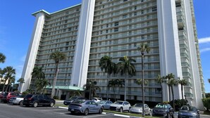 Hotel de varios pisos con fachada blanca y techo verde, que cuenta con un estacionamiento con varios coches y palmeras.