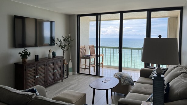 Amplia sala de estar con televisor grande, mueble de madera, pequeña mesa y sofá. Balcón con vista al océano.