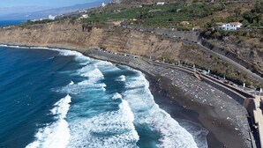 A coastal road with a beach on the right, a rocky cliff on the left, and houses in the background. - Home2Book Atlantic View Rustic Villa (Los Realejos)
