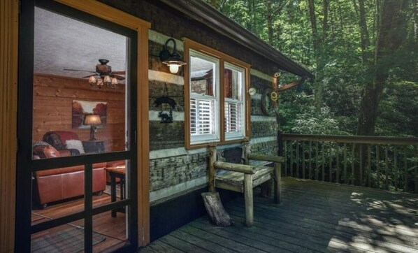 Une cabane dotée d’une terrasse en bois, d’un intérieur chaleureux avec un canapé en cuir et une table, et d’une fenêtre donnant sur la forêt.