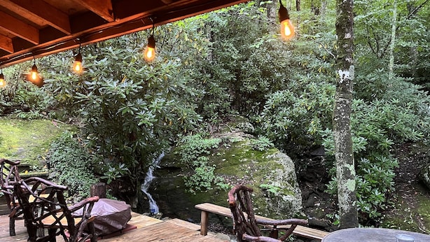 An outdoor wooden deck with rustic chairs and a round table, surrounded by lush greenery and a small waterfall. - PEACOCK RIDGE Great location, minutes from Blowing Rock and Appalachian Ski Mtn! (Blowing Rock)