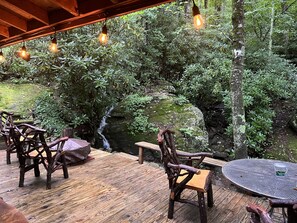 Une terrasse extérieure en bois, agrémentée de chaises rustiques et d’une table ronde, entourée d’une végétation luxuriante et d’une petite cascade.