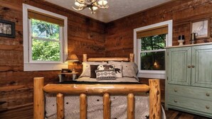 A rustic wooden room with a bed featuring a wooden headboard, a green cabinet, and two windows with views of greenery. - PEACOCK RIDGE Great location, minutes from Blowing Rock and Appalachian Ski Mtn! (Blowing Rock)
