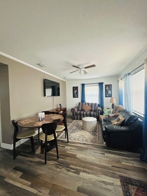 Un salon comprenant un coin repas, un ventilateur de plafond, un téléviseur à écran plat et un canapé agrémenté de coussins à motifs.