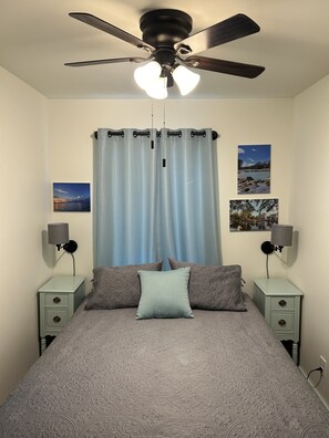 Une chambre à coucher comprenant un lit, deux tables de chevet, un ventilateur de plafond et une fenêtre avec des rideaux.