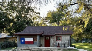 Une petite maison avec le drapeau du Texas peint sur le mur latéral, une porte rouge et un chemin de pierres menant à l’entrée.