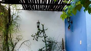Un patio con dos sillas blancas de metal y una pequeña mesa azul, rodeado de plantas y un muro azul.