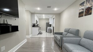 A modern living room with a grey sofa, a flat-screen TV mounted on the wall, and a kitchen in the background. - Adorable 2 bedroom guest house! Minutes from downtown (Houston)