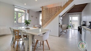 A modern kitchen with a wooden staircase, a dining area with white chairs, and a view of the outdoors. - La Maison aux Portes des Ecluses (HEDE BAZOUGES)