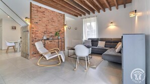 A modern living room with a brick accent wall, a grey sofa, white chairs, and a dining area with a table and chairs. - La Maison aux Portes des Ecluses (HEDE BAZOUGES)