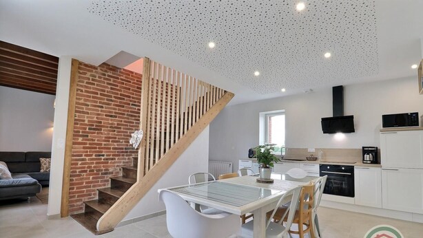 A modern kitchen with white cabinets, a black microwave, and a coffee maker. There's a dining area with a white table and chairs, and a staircase with a wooden railing leading to an upper level. - La Maison aux Portes des Ecluses (HEDE BAZOUGES)