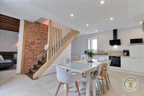 A modern kitchen with white cabinets, a black microwave, and a coffee maker. There's a dining area with a white table and chairs, and a staircase with a wooden railing leading to an upper level.