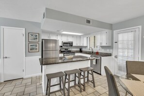 A modern kitchen with white cabinets, a stainless steel refrigerator, a microwave, and a gas stove. There are three stools at the island and a window with blinds.