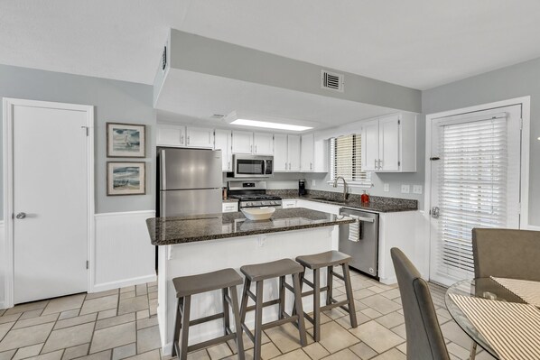 A modern kitchen with white cabinets, a stainless steel refrigerator, a microwave, and a gas stove. There are three stools at the island and a window with blinds.