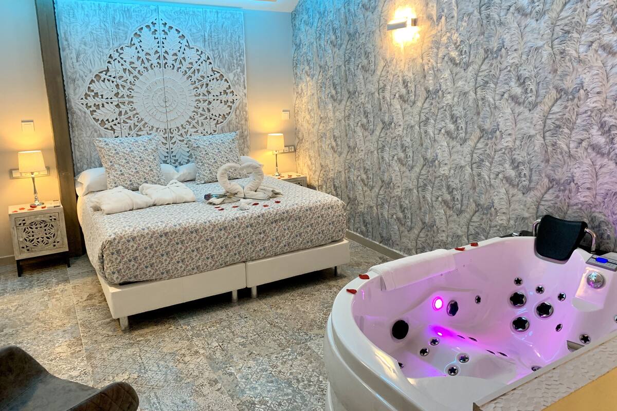 A bedroom with a bed, a decorative headboard, a nightstand with a lamp, a bathroom with a bathtub, and a wall with a textured pattern.