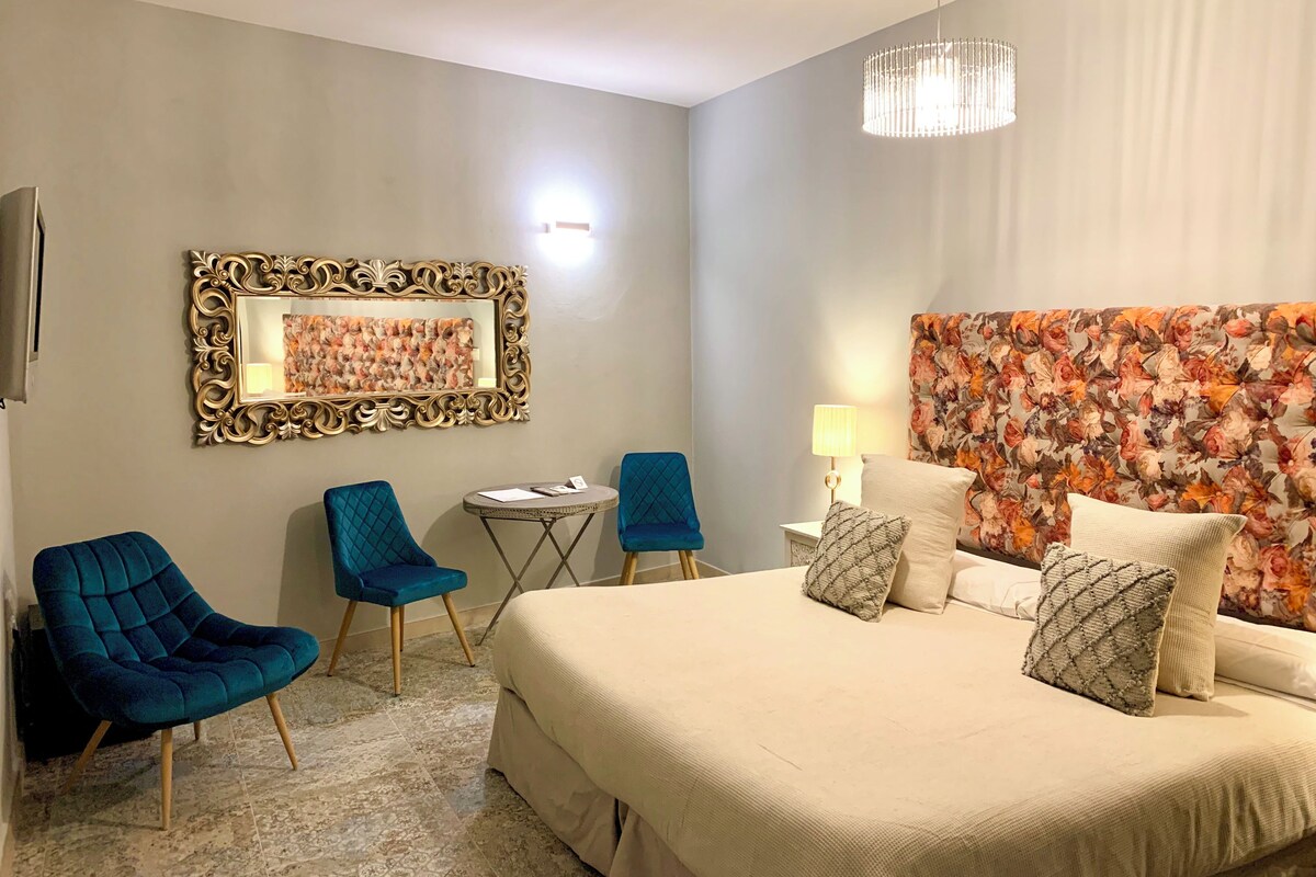 Quarto de hotel com uma cama grande, duas poltronas azuis, uma mesa redonda pequena, um abajur e uma cabeceira estampada com flores.