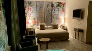 A hotel room with a large bed, a sitting area with a chair and a small table, a bathtub with lights, a chandelier, and a television on the wall.