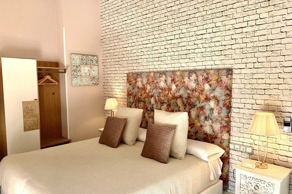 A bedroom with a brick-patterned headboard, a bed with multiple pillows, a bedside table with a lamp, and a wardrobe with a mirror.