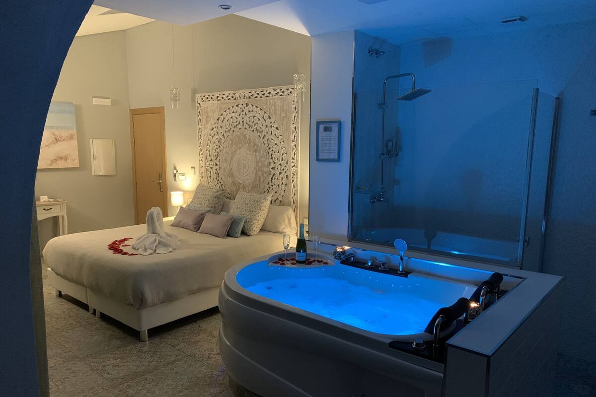 A bedroom with a large bathtub, a bed with decorative pillows, and a shower area.