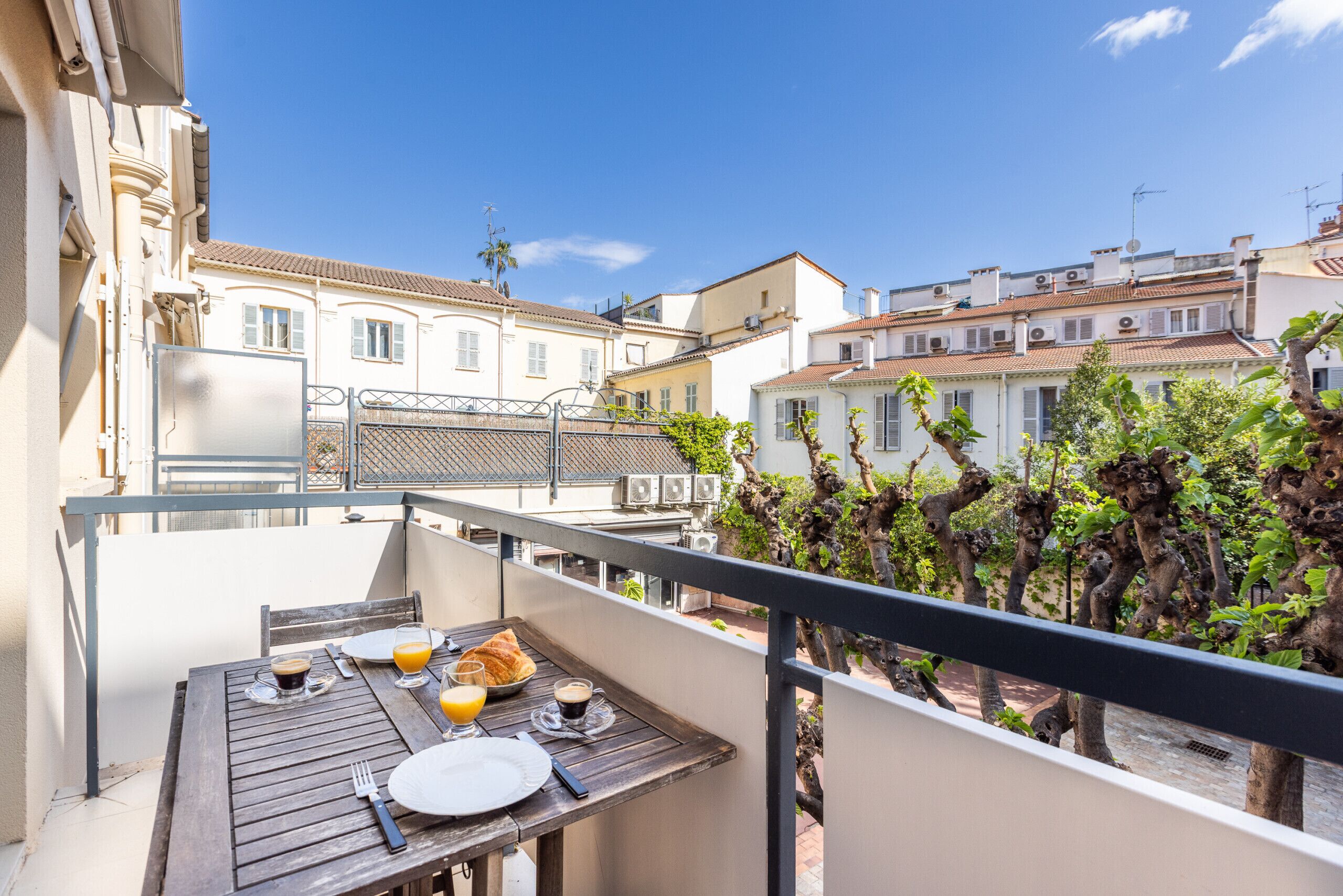 A balcony with a wooden table set for breakfast, overlooking a cityscape with buildings and trees.