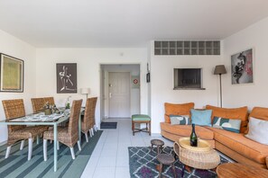 Un salon avec un canapé, une table à manger avec des chaises, une petite table sur laquelle se trouve une bouteille, et une porte qui donne sur une autre pièce.