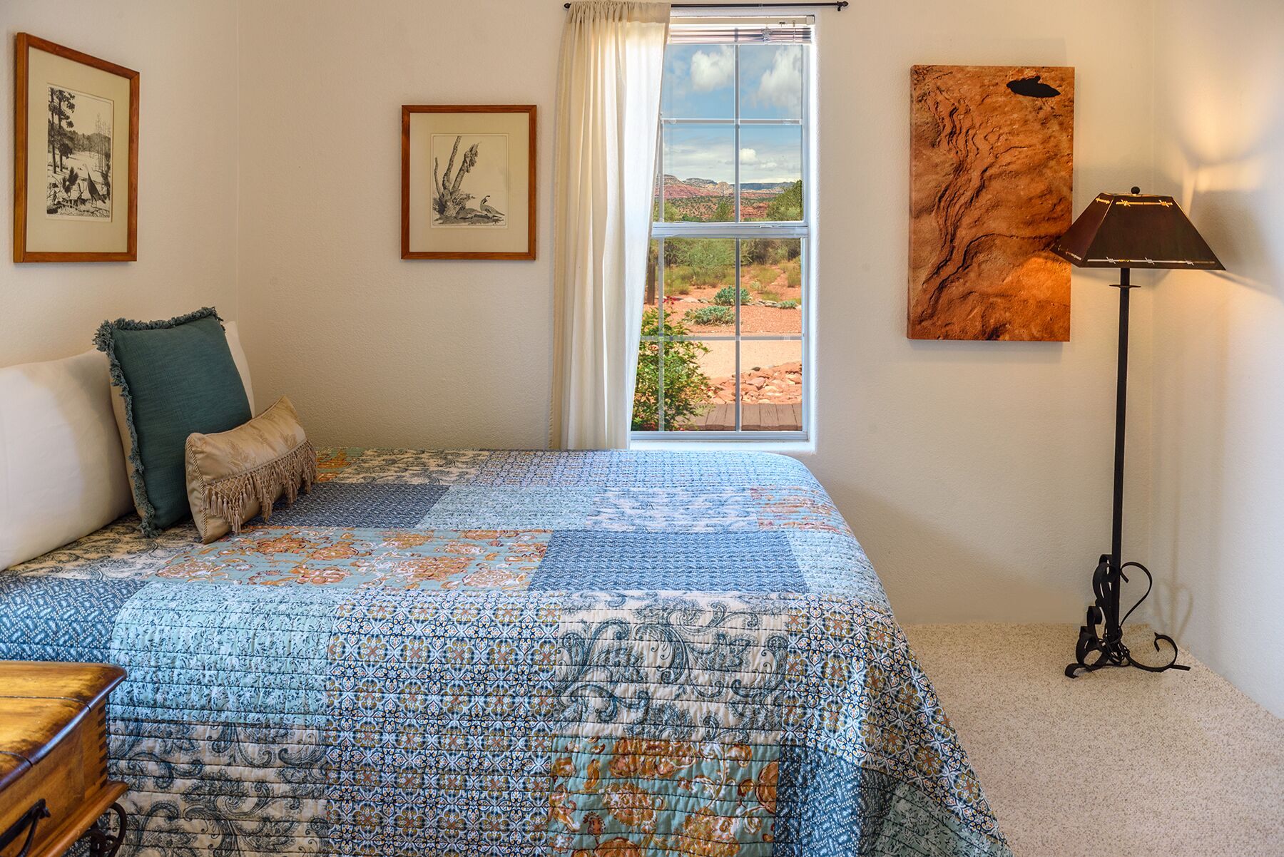 A bedroom with a large bed, a window with a view, a lamp, and two framed pictures on the wall.