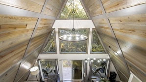 A high-ceilinged room with wooden walls and large windows, a central chandelier, and a dining area with a table and chairs.