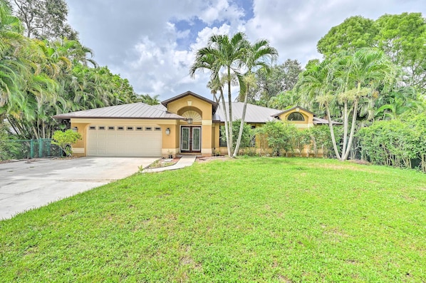A two-story house with a garage, surrounded by lush greenery and palm trees.