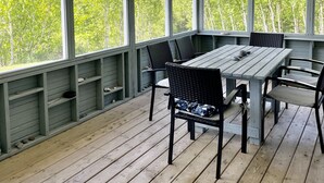 Un espacio al aire libre cubierto con mesa y sillas, ventanas amplias y vista a árboles.