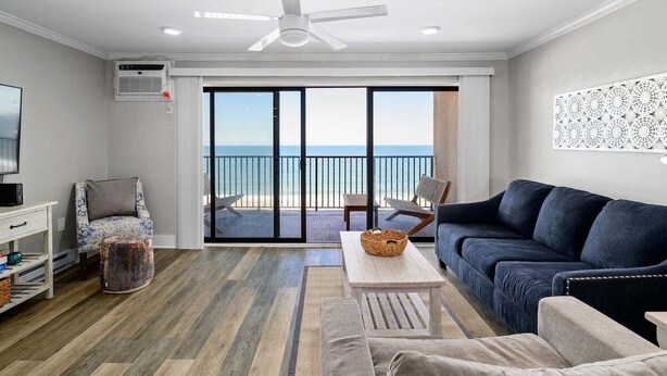 A living room with a blue sofa, a white coffee table, a patterned armchair, and a television on a white stand. - Oceanfront 1 Bedroom, Balcony, Sitting Room, Pool (Ocean City)