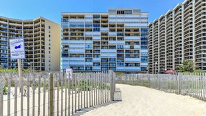 A beachfront property with a wooden fence, a sign indicating a beach access point, and high-rise buildings in the background. - Oceanfront 1 Bedroom, Balcony, Sitting Room, Pool (Ocean City)