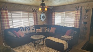 Ein gemütliches Wohnzimmer mit einem Ecksofa, einem Couchtisch und einem Teppich. An der Wand hängt eine Uhr und es gibt einen Deckenventilator.