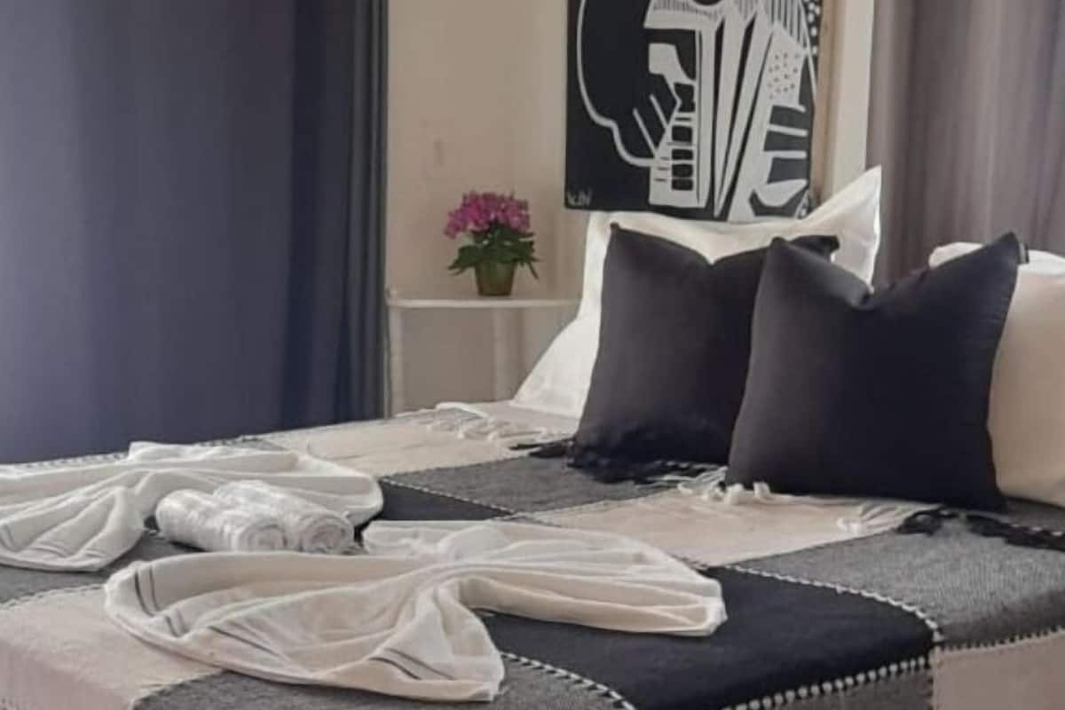 A neatly made bed with a black and white checkered bedspread, two black pillows, and a white pillow. There is a small table with a potted plant in the background.