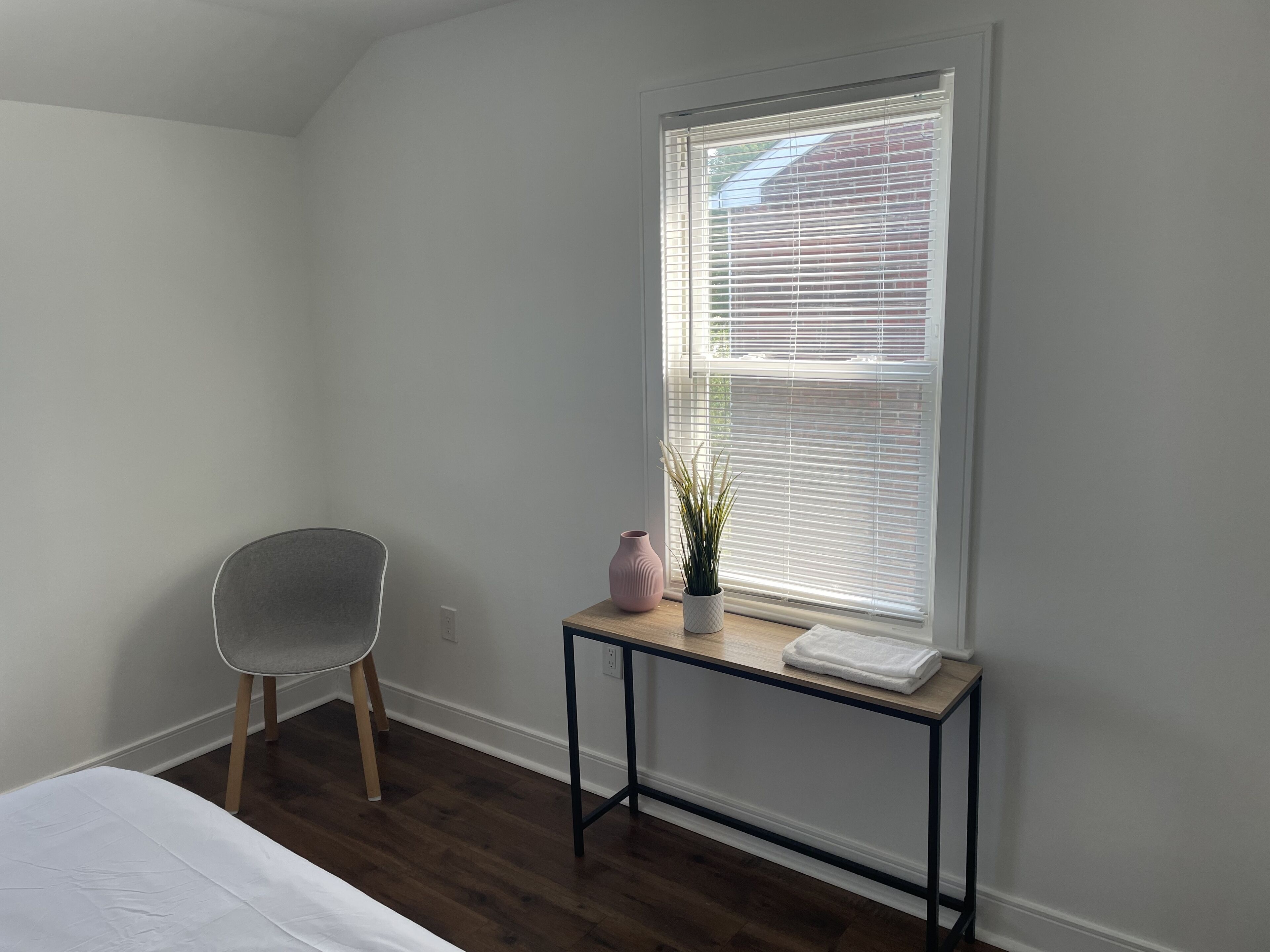 Ein Schlafzimmer mit einem Bett, einem Sessel, einem kleinen Tisch mit Vase und Pflanze sowie einem Fenster mit Jalousien.