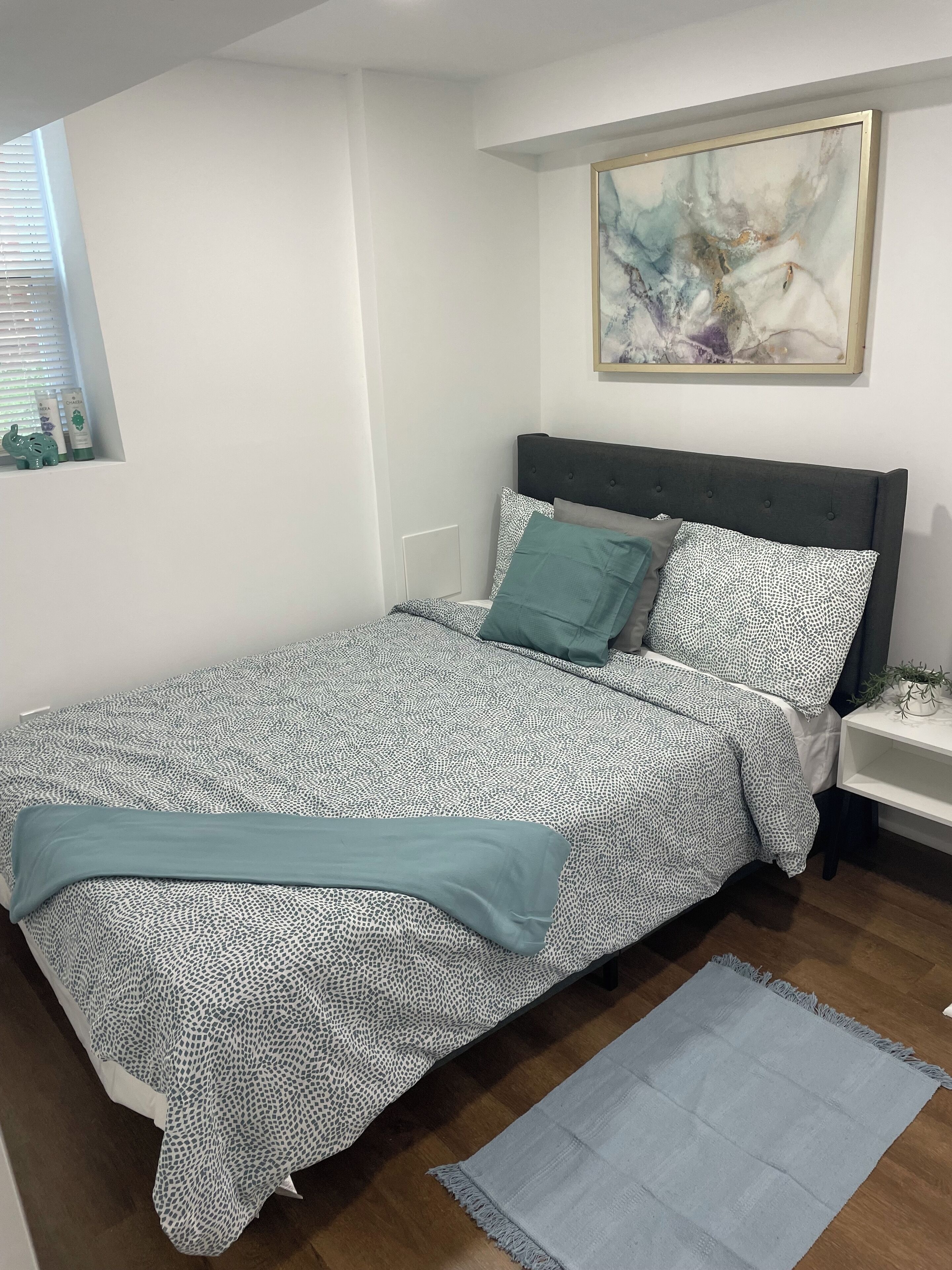 A neatly made bed with patterned bedding, a bedside table with a lamp, and a wall-mounted abstract painting.