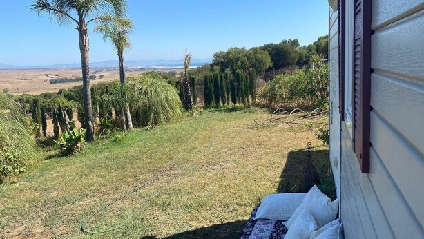 A backyard with a view of distant hills, a palm tree, and a fence. - Cozy tiny house in nature, 5 mins from Beach | With sea view & high speed WiFi (Tarifa)