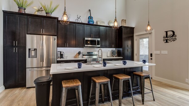 Une cuisine moderne avec des meubles de couleur sombre, des appareils en acier inoxydable, un îlot central avec des tabourets, des luminaires suspendus et des objets décoratifs sur le plan de travail.